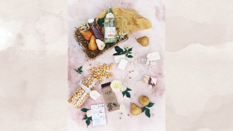 Wedding welcome bag snacks with fruit, popcorn, drinks, and small favors on a pink table